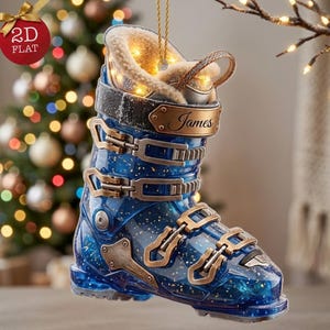 May include: A blue translucent ski boot ornament featuring gold-toned buckles, a beige faux-fur lining, and a gold nameplate engraved with the name James. The boot includes an integrated string for hanging and internal lights that illuminate the interior.