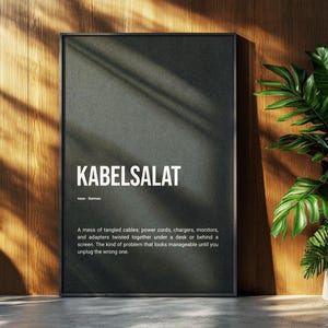 Könnte beinhalten: Ein rechteckiges Wandbild mit dunkelgrauem Hintergrund und dem weißen Text KABELSALAT, noun / German, gefolgt von einer Definition, die ein Durcheinander von verhedderten Kabeln, Netzkabeln, Ladegeräten und Adaptern unter einem Schreibtisch beschreibt.