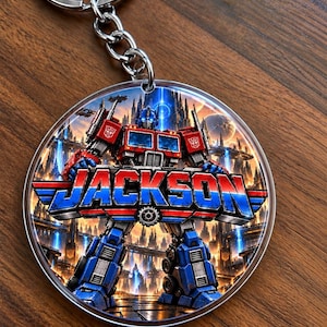May include: A circular keychain featuring a graphic of a red and blue robot character standing in a futuristic city. The robot holds a large, stylized blue and red banner with the name JACKSON printed in bold, blocky, metallic-textured letters.
