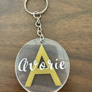 May include: A circular clear acrylic keychain featuring a large gold letter A in the center with the name Avorie printed in white script lettering across the front. The keychain is attached to a silver metal split ring by a short chain.