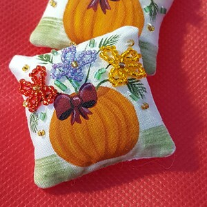 May include: Two small, square decorative pillows with a pumpkin design. Each pillow features a painted orange pumpkin, a burgundy bow, and beaded flowers in red, purple, and yellow. The pillows are on a red background.