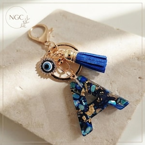 Peut inclure: Un porte-clés doré avec une breloque en forme de lettre A en résine bleu foncé et bleu sarcelle incrustée de feuilles d'or. Il comprend un pompon en daim bleu et une petite breloque ronde représentant un œil bleu et blanc entouré de strass.