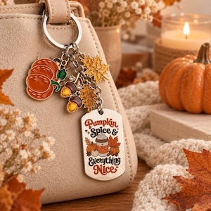 Pode incluir: Um porta-chaves de metal prateado preso a uma mala bege, com berloques de uma abóbora laranja, duas bolotas, uma folha de ácer dourada e uma etiqueta retangular com o texto Pumpkin Spice & Everything Nice e uma chávena de café.