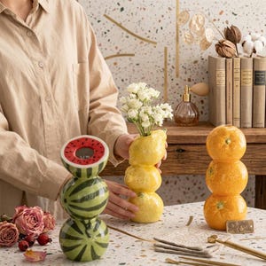Può includere: Tre vasi in ceramica a forma di frutta impilata: un'anguria verde, un limone giallo e un agrume arancione. Il vaso a forma di limone contiene un piccolo bouquet di fiori bianchi. Ogni vaso presenta una finitura lucida e texture realistiche.