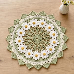 May include: A round crochet doily featuring a central sage green lattice pattern surrounded by a ring of white daisy motifs with yellow centers. The edge is finished with a scalloped border in cream and sage green yarn, creating a floral design.