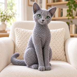 Peut inclure: Une figurine de chat crochetée grise faite à la main avec une surface en fil texturé, des oreilles pointues et des yeux vert vif. Le chat est représenté en position assise avec la queue recourbée sur le côté, montrant des points détaillés.