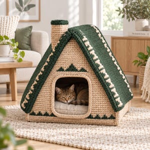 Puede incluir: Una casa para mascotas de forma triangular hecha de hilo de ganchillo beige y verde oscuro. La estructura cuenta con una chimenea, un techo puntiagudo con un patrón de zigzag blanco y un diseño de montañas sobre la entrada donde descansa un gato.