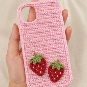 May include: A light pink crochet phone case featuring two small, red strawberry appliqués with green leaves and white embroidered seeds. The case has a rectangular shape with a circular cutout at the top left corner for a camera lens.