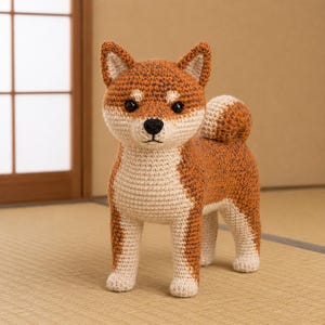以下が含まれることがあります： 茶色とクリーム色の糸で編まれた、かぎ針編みの柴犬のぬいぐるみ。尖った耳、黒い鼻、暗い色の目、巻いた尻尾が特徴で、胸と腹部がクリーム色をしており、リアルな立ち姿で表現されています。
