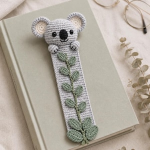 May include: A gray crocheted koala bookmark featuring a long rectangular body with a green vine and leaf pattern embroidered down the center. The koala head has round ears with cream-colored centers, black eyes, and a black stitched nose and mouth.