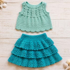May include: A two-piece crochet outfit consisting of a sleeveless mint green top with a scalloped neckline and hem, and a tiered teal skirt with a ruffled design. The set features intricate openwork stitching throughout both pieces.