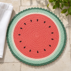May include: A round, crocheted placemat designed to resemble a watermelon slice. The center is bright coral-red with scattered black teardrop-shaped stitches representing seeds, surrounded by a thin white ring and a wide, scalloped dark green border.