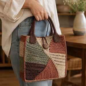 Può includere: Una borsa all'uncinetto di forma quadrata con un design patchwork nei toni del verde oliva, terracotta, marrone scuro e crema. La borsa presenta due manici all'uncinetto marrone scuro e una chiusura centrale a bottone circolare.