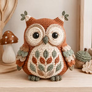 Può includere: Un peluche a forma di gufo realizzato all'uncinetto con corpo color ruggine, dischi facciali color crema e grandi occhi neri. Il petto è decorato con un motivo di piccole foglie ricamate nei toni del verde salvia e ruggine.