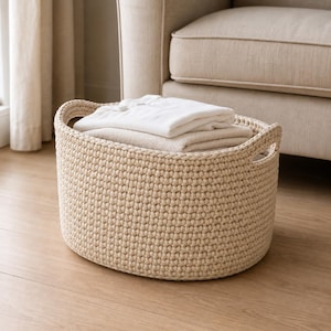 May include: An oval-shaped storage basket crafted from thick, cream-coloured cord with a textured, crocheted stitch pattern and integrated side handles. The basket is filled with folded white and beige fabric items, creating a neutral storage solution.
