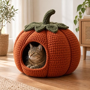 Puede incluir: Una cama redonda para mascotas, tejida a ganchillo en color naranja con forma de calabaza, que presenta un tallo y hojas de color verde oscuro en la parte superior. Un gato atigrado marrón con ojos verdes descansa dentro de la abertura circular.