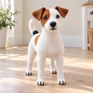 Może przedstawiać: Szydełkowana figurka psa rasy Jack Russell Terrier wykonana z białej, brązowej i czarnej włóczki. Pies ma białe ciało z brązowymi łatami na uszach i grzbiecie, czarny nos oraz ciemne oczy. Tekstura włóczki jest widoczna na całej figurce.