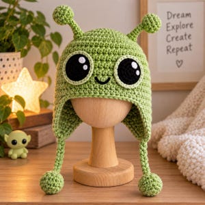 May include: A light green crocheted hat featuring two antennae with round tips, large black circular eyes with white highlights, and a small black embroidered smile. The hat includes two long braided earflaps ending in small green crocheted spheres.