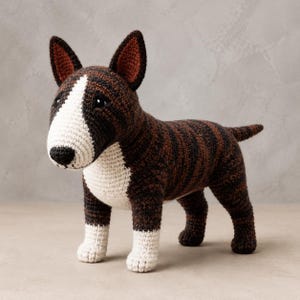 May include: A crochet bull terrier dog figurine featuring a dark brown and black brindle patterned body with a white chest, white lower legs, and a white face patch. The dog has pointed ears with dark interiors and a black stitched nose.