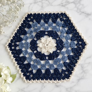 May include: A hexagonal crochet doily featuring a central cream-colored flower motif surrounded by concentric layers of navy blue and light blue yarn. The piece is finished with a delicate cream-colored scalloped border along all six edges.