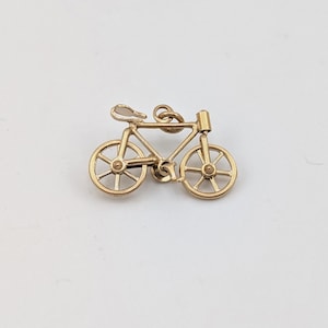 May include: A gold-toned metal bicycle charm featuring a detailed frame, two spoked wheels, a seat, and a small jump ring attached to the top bar for hanging. The design captures the literal shape of a bicycle with a central pedal and handlebar.