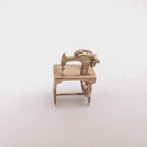 May include: A gold-colored metal charm shaped like a vintage sewing machine. The charm features a flat top surface, a vertical arm with a needle bar, a side-mounted wheel with spokes, and a small circular jump ring attached to the top for hanging.
