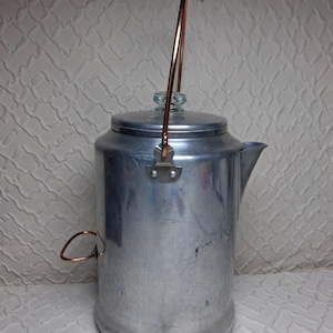 Puede incluir: Una cafetera percoladora cilíndrica y alta de aluminio con acabado metálico pulido, un asa de alambre color cobre, un mango lateral a juego color cobre, una tapa ajustada y un pequeño pomo de vidrio transparente en la parte superior.