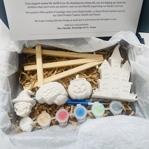 May include: A craft kit in a box containing a wooden easel, a white plaster skull, a brain, a pumpkin, and a haunted house figure. Included are two paintbrushes and a strip of six paint pots with white, yellow, red, blue, green, and black paint.