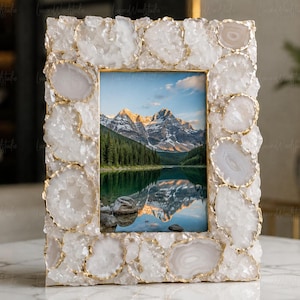 May include: A rectangular picture frame featuring a border composed of numerous white and translucent agate stone slices with gold-toned metallic edges. The frame holds a photograph of a snow-capped mountain range reflected in a calm lake.