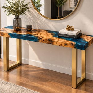May include: A rectangular console table featuring a natural wood burl slab with blue resin inlay, supported by two gold-toned metal U-shaped legs. The table top has a thickness of 35 mm. A glass vase, a candle, and two books are placed on the surface.