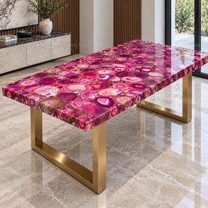 May include: A rectangular dining table featuring a polished tabletop composed of numerous vibrant pink and magenta agate slices with a glossy finish, supported by two sturdy, brushed gold-toned metal U-shaped legs on a reflective floor surface.