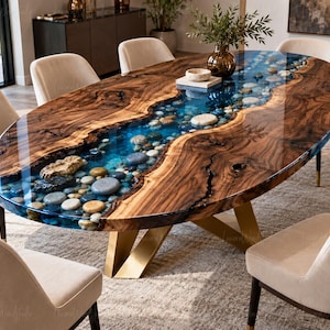 Può includere: Un tavolo da pranzo ovale con un piano in legno a bordo naturale e un design a fiume centrale realizzato in resina blu traslucida riempita di ciottoli lisci. Il tavolo è sostenuto da una base in metallo dorato ed è circondato da sedie beige imbottite.