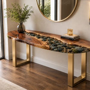 Op de afbeelding: Een consoletafel met een houten blad met natuurlijke randen en een centrale rivierachtige inleg van gladde, veelkleurige rivierstenen ingebed in heldere hars. De tafel wordt ondersteund door twee rechthoekige poten van geborsteld goudkleurig metaal.