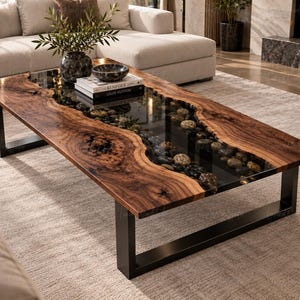 Puede incluir: Una mesa de centro rectangular con dos tablones de madera de borde natural y una incrustación central de vidrio rellena de piedras de río lisas. La mesa se apoya en patas de metal negro en forma de U y sostiene libros Kinfolk y Louis Vuitton.