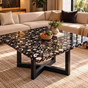 May include: A square coffee table featuring a polished black surface inlaid with numerous circular, white and grey stone slices. The table is supported by a black metal base with a cross-shaped design. A small woven basket of fruit sits on top.