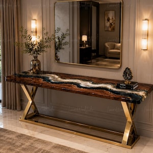 May include: A long console table featuring a live-edge dark wood slab with a central river-like inlay of swirling blue, white, and black resin. The table is supported by a polished gold-tone metal X-frame base and sits against a wall.