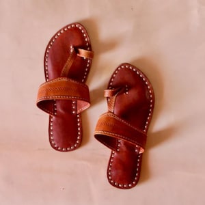 May include: A pair of brown leather sandals featuring a toe loop design and a wide horizontal strap across the midfoot. The edges of the soles are detailed with visible white stitching, and the leather has a smooth, polished finish throughout.