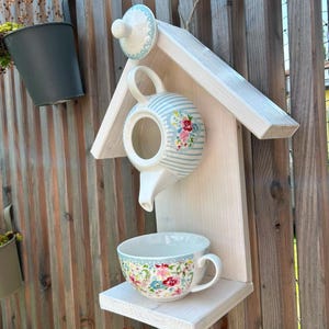 Könnte beinhalten: Eine weiße Wandhalterung aus Holz in Vogelhausform mit einer Keramik-Teekanne mit hellblauen Streifen und Blumenmuster in der Mitte, einer passenden Blumentasse auf der unteren Ablage und einem Teekannendeckel an der Spitze.