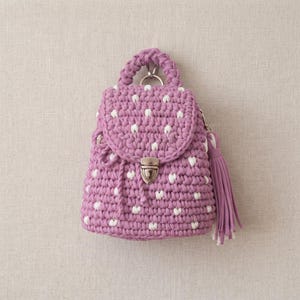 Puede incluir: Una pequeña mochila de ganchillo hecha a mano en color malva polvoriento con un patrón repetitivo de diminutos puntos blancos. El bolso cuenta con un asa superior, un cierre de solapa con hebilla de metal plateado y una borla malva a juego en el lateral.