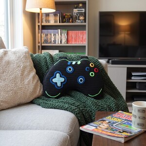 May include: A black crochet pillow shaped like a video game controller. The pillow features a light blue directional pad, two blue joysticks, and four colored action buttons in yellow, blue, green, and red. A thin lime green line accents the bottom.