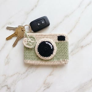 May include: A rectangular crochet pouch shaped like a camera, featuring sage green and cream yarn, a prominent black circular lens detail, and a small leaf-shaped charm. A gold-toned metal keyring is attached, holding a black car key fob and keys.