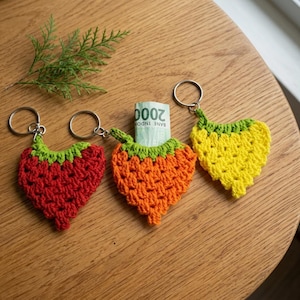 Puede incluir: Tres llaveros de ganchillo en forma de corazón en rojo, naranja y amarillo, cada uno con una parte superior verde y una anilla de metal plateado. El llavero naranja contiene un billete de 2000 rupias indonesias enrollado en su abertura.