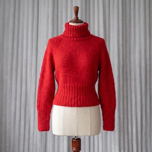 Könnte beinhalten: Ein leuchtend roter Langarmpullover mit einem dicken Rollkragen und einer gerippten Strickstruktur an den Ärmelbündchen und am Saum. Das Kleidungsstück wird auf einer Schneiderpuppe präsentiert und zeigt eine taillierte Silhouette aus Grobstrick.