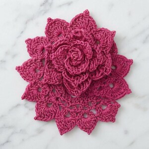 May include: A vibrant magenta crochet flower applique featuring layered, scalloped petals arranged in a circular, star-like pattern. The intricate needlework creates a textured, three-dimensional floral design with a dense, spiraled center.
