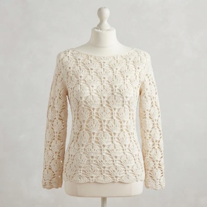 May include: A long-sleeved sweater in an off-white cream color, featuring an intricate, repeating openwork crochet pattern throughout the body and sleeves. The garment has a wide, rounded neckline and a scalloped hemline along the bottom edge.