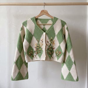 May include: A cropped crochet cardigan featuring a green and cream argyle diamond pattern. The garment has long bell sleeves, a V-neckline, and three white buttons down the front. Delicate floral embroidery in yellow, orange, and green accents the front.