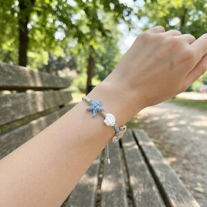 May include: A braided bracelet featuring a light blue crocheted starfish, a white crocheted seashell, and a beige crocheted seahorse. The bracelet is constructed from light blue and beige cord, with the charms arranged along the band.