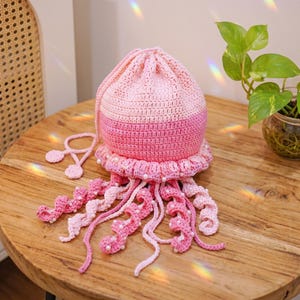 May include: A handmade crochet jellyfish bag in shades of light and dark pink. The rounded body features a drawstring closure with two circular pulls, while the base is adorned with dangling, spiraled tentacles accented with small white beads.
