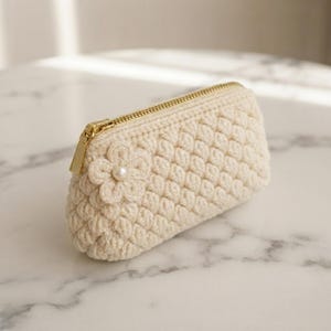 May include: A cream-colored crochet pouch featuring a textured popcorn stitch pattern. The pouch has a gold-tone metal zipper closure and is decorated with a single crocheted flower accent centered with a small white pearl bead.