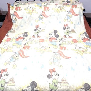 May include: A cream-colored bedspread featuring a repeating pattern of Disney characters including Mickey Mouse, Minnie Mouse, Donald Duck, and Pluto. The print includes images of birthday cakes, balloons, and flags labeled Magic Kingdom.